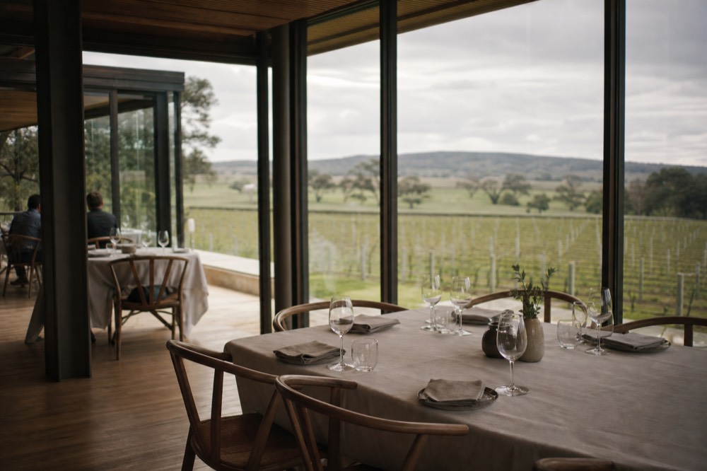 A refined winery dining room with floor-to-ceiling windows onto vineyard rows, linen-set tables and timber floors, soft natural light, no branding, menus or people.