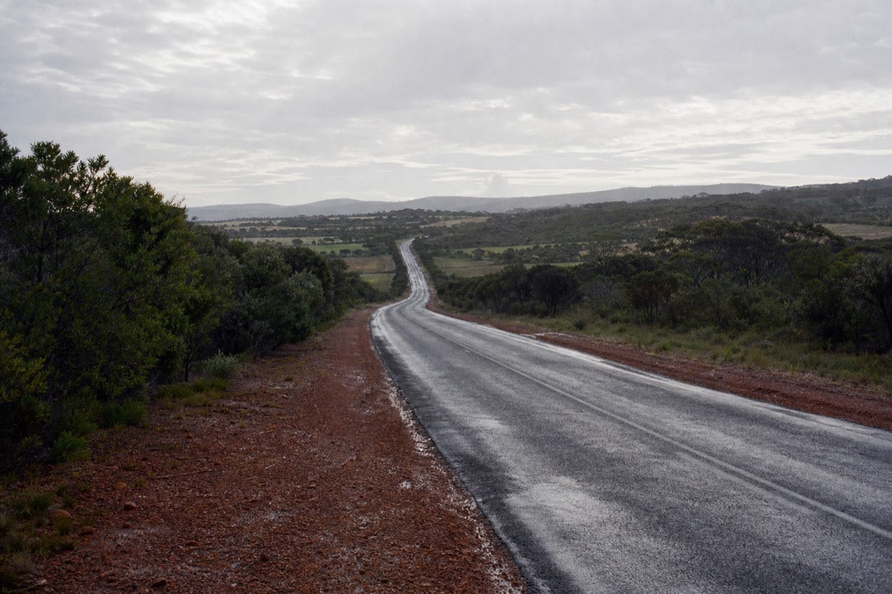 An open highway running south through low coastal bushland and farmland under a bright sky, distant hills ahead, empty road, no signage, vehicles or people.