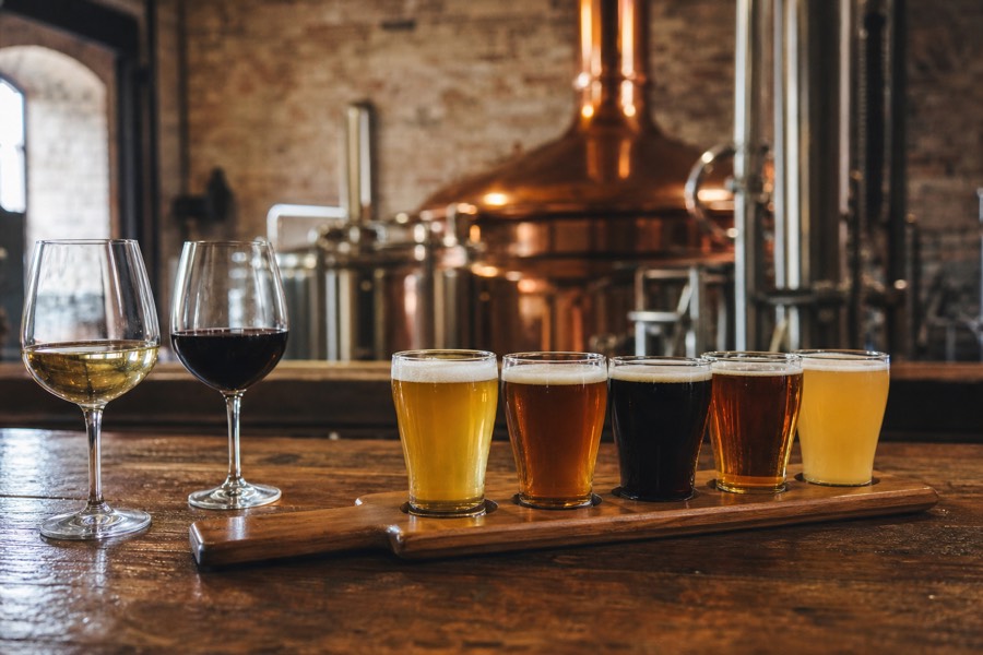 A tasting paddle of craft beer alongside wine glasses on a brewery bar, distillery copper still softly out of focus behind