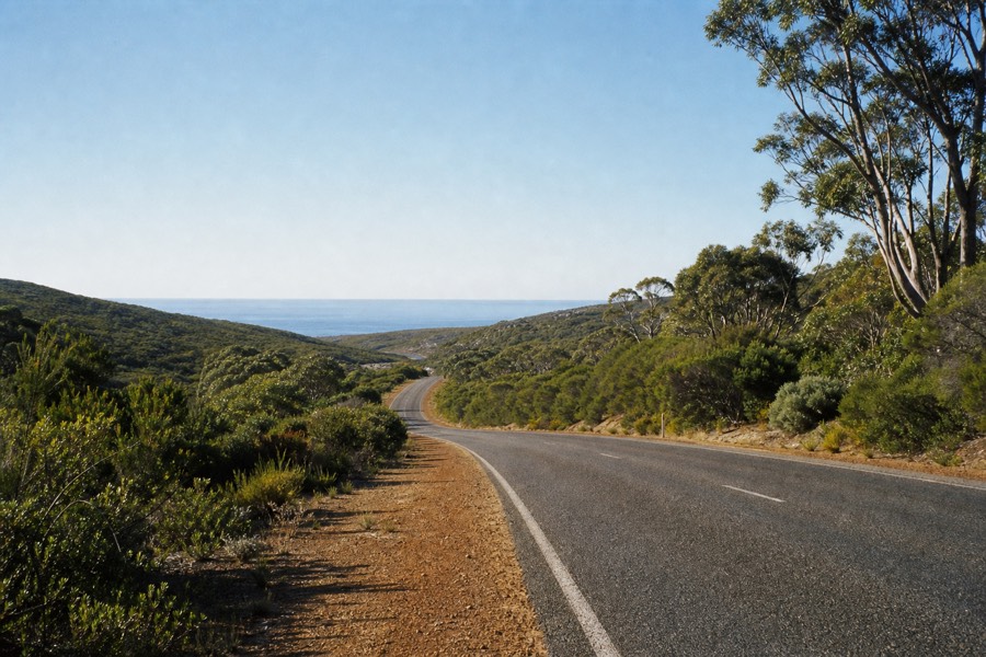 A long coastal road curving south toward the Margaret River region, ocean and low forest either side under clear morning light