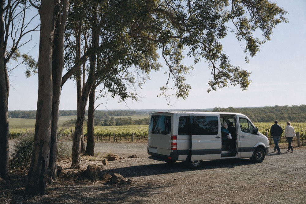 A small tour van parked on gravel beside vineyard rows, doors open, a wide green valley beyond, clear afternoon light, no branding or number plates visible.