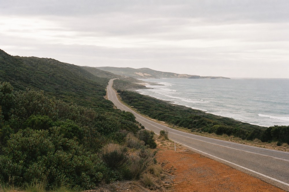 A long coastal highway seen from a low rise, ocean on one side and bush on the other, soft morning light, no vehicles or signage.