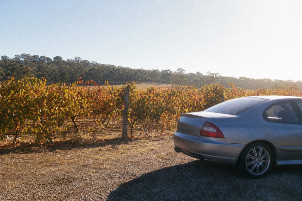 A car parked on gravel in full sun beside a vineyard fence, heat haze shimmering above the bonnet, dry grass and bright sky, no people or animals visible.