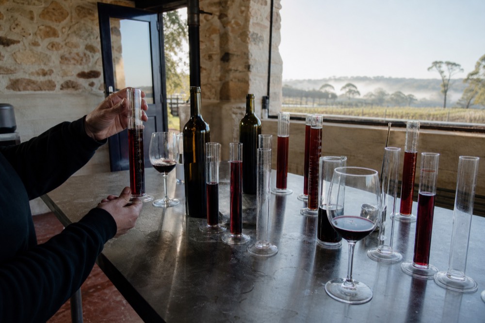 Hands at a blending bench with measuring cylinders and unlabelled wine samples, soft daylight, no faces or branding.