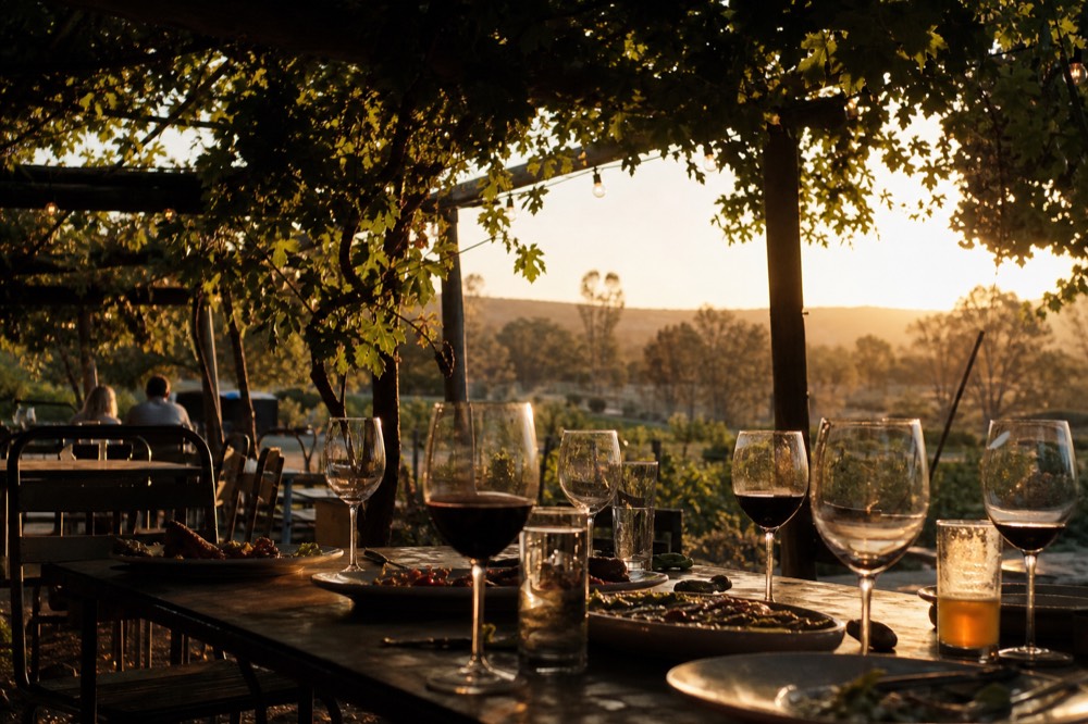 A group table under a pergola set with a mix of wine glasses and beer glasses beside shared plates, greenery overhead, warm light, no diners visible.