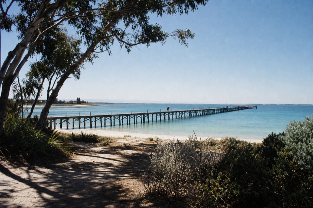 A long timber jetty reaching far out over calm turquoise water under a clear sky, pale sand and low coastal scrub in the foreground, no people or branding.