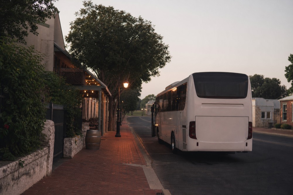 A quiet city street at dawn with a waiting touring vehicle at the kerb, warm streetlight and a pale sky above low buildings, no passengers, plates or signage visible.