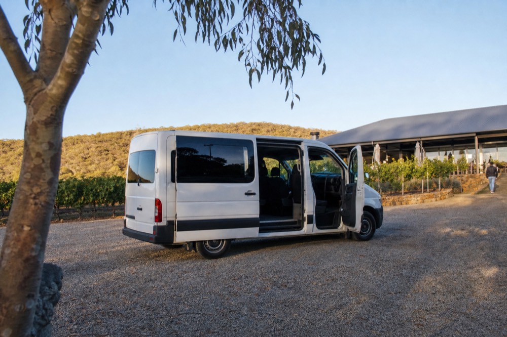 A small tour van parked on gravel outside a vineyard restaurant at midday, doors open, vine rows and a low hill behind, clear bright light, no branding or number plates visible.
