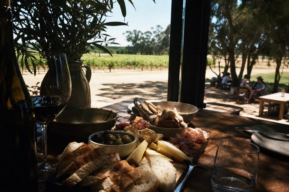 A shared platter of local cheese, bread, olives and cured meats down the centre of a table beside a glass of red wine, a vineyard visible through a window, warm daylight, no logos or text.