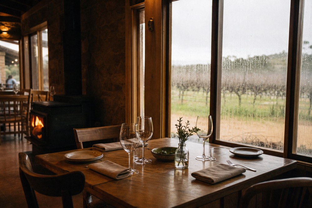 A cosy winery dining room with a lit fireplace and a table set for lunch, rain-streaked windows looking onto bare-canopy winter vines, warm interior light, unbranded, no people.