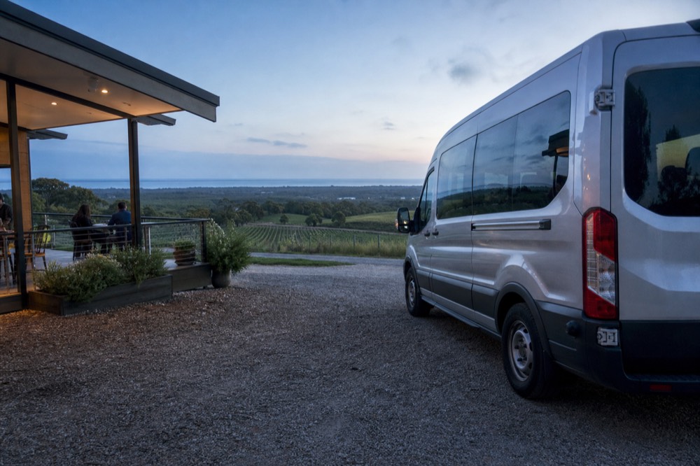 A small tour van parked on gravel beside vineyard rows with a wide green valley beyond and a hint of coast on the horizon, clear afternoon light, no branding or number plates visible.