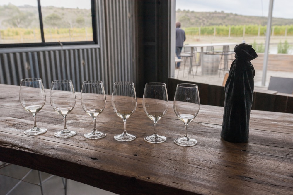 A row of unlabelled wine glasses set up for a blind tasting on a rustic timber bench, a covered bottle beside them, soft daylight through a shed window, no branding or text, no people.